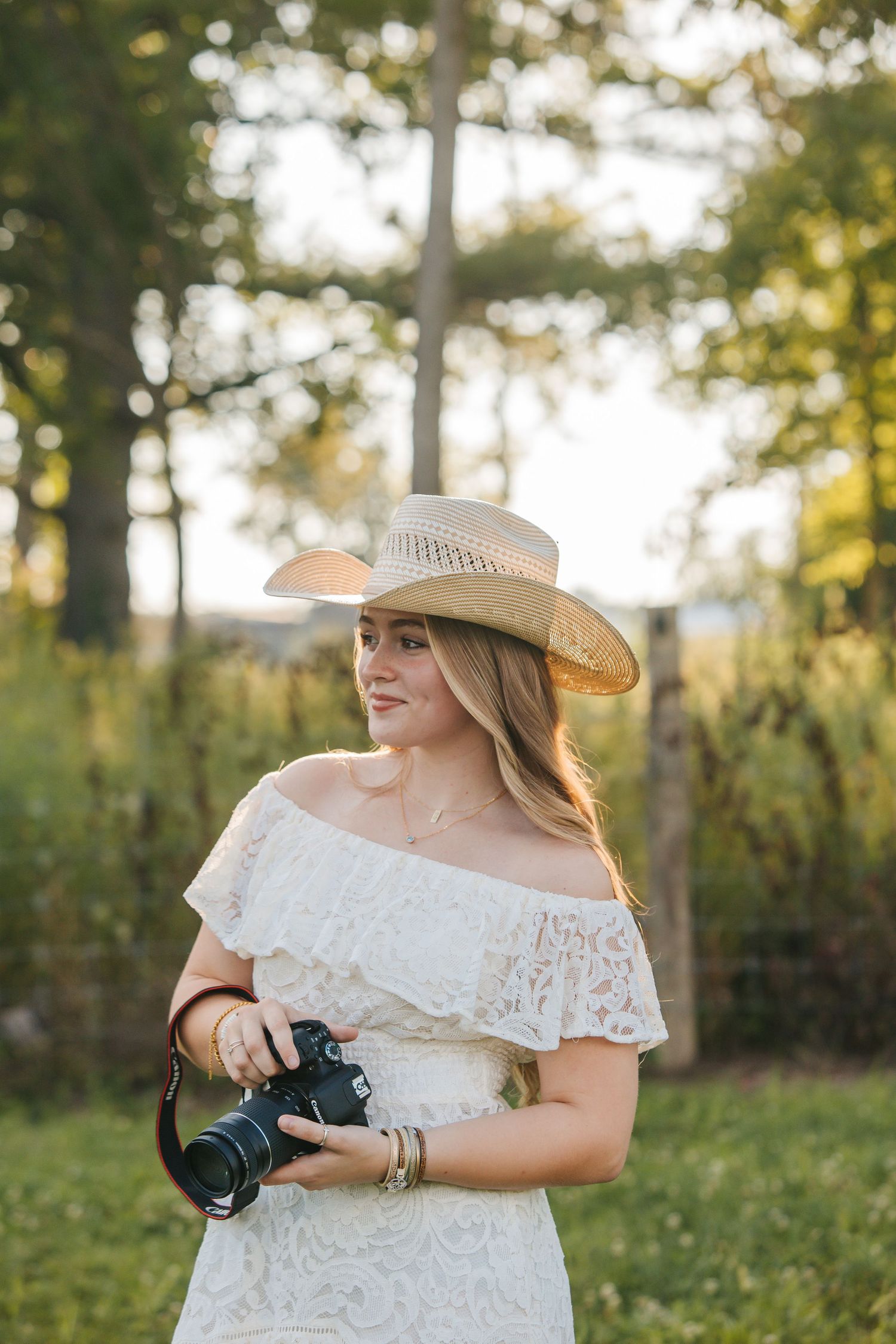 A photographer in a white lace dress and sun hat poses with a camera in a sunlit outdoor setting.