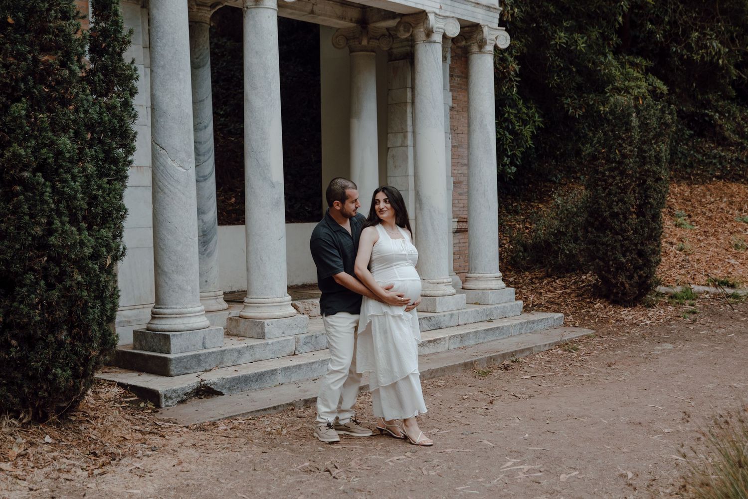 A couple stands in a white columned gazebo surrounded by autumn foliage in a romantic outdoor setting.