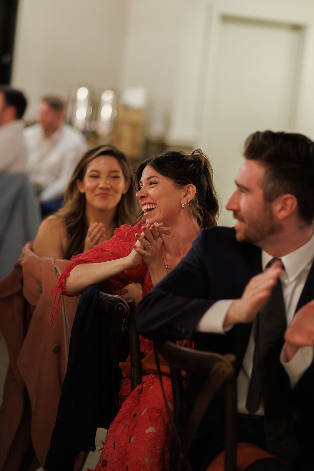 People laughing together at an indoor event, with one person in a red dress covering their mouth while laughing.