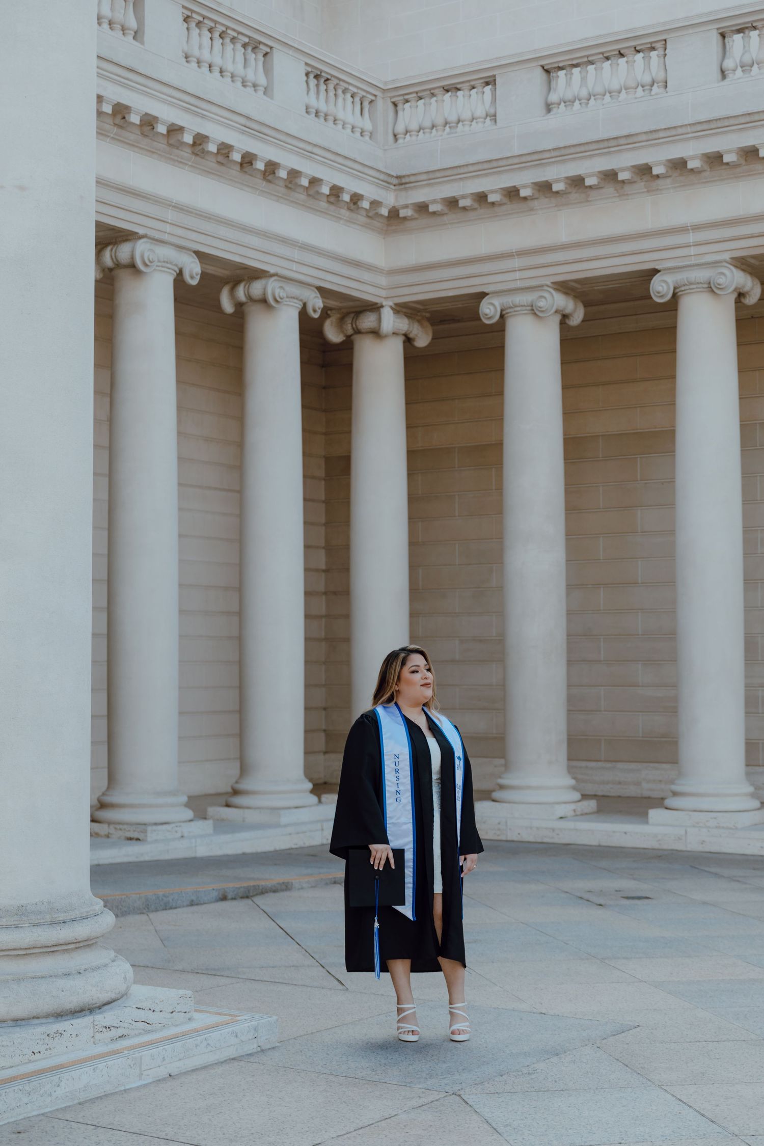 A person in a black dress stands between classical white columns in an elegant architectural setting.