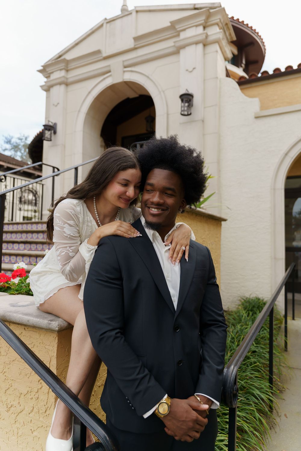 A couple shares a romantic moment on outdoor steps of a Spanish-style building with cream walls and wrought iron railings.