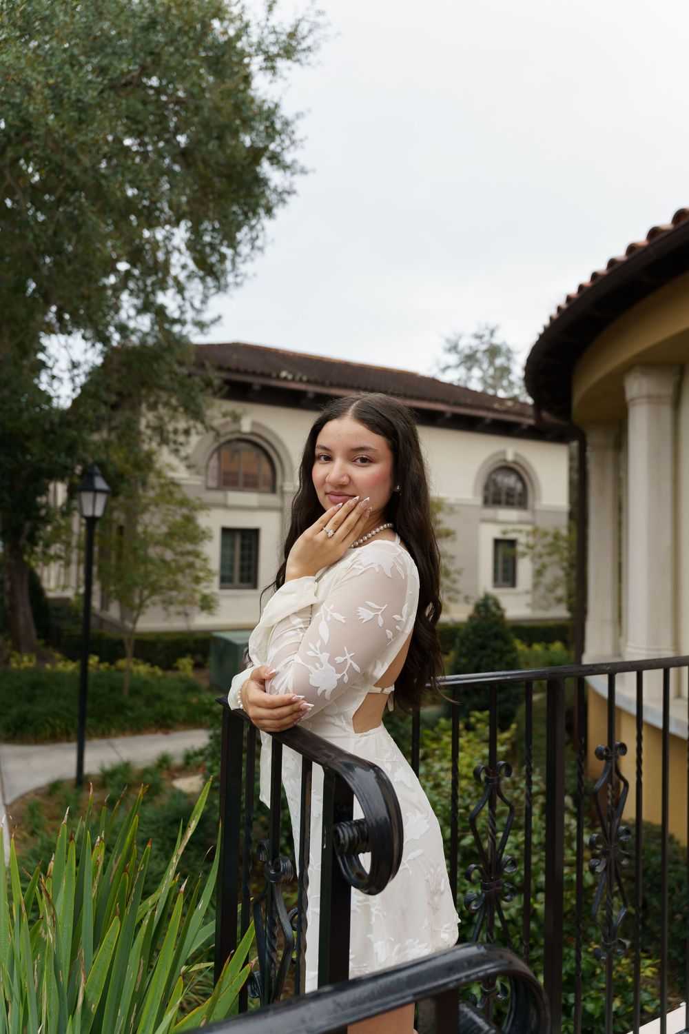 Woman in white lace dress poses on a railing at an elegant Spanish-style building.
