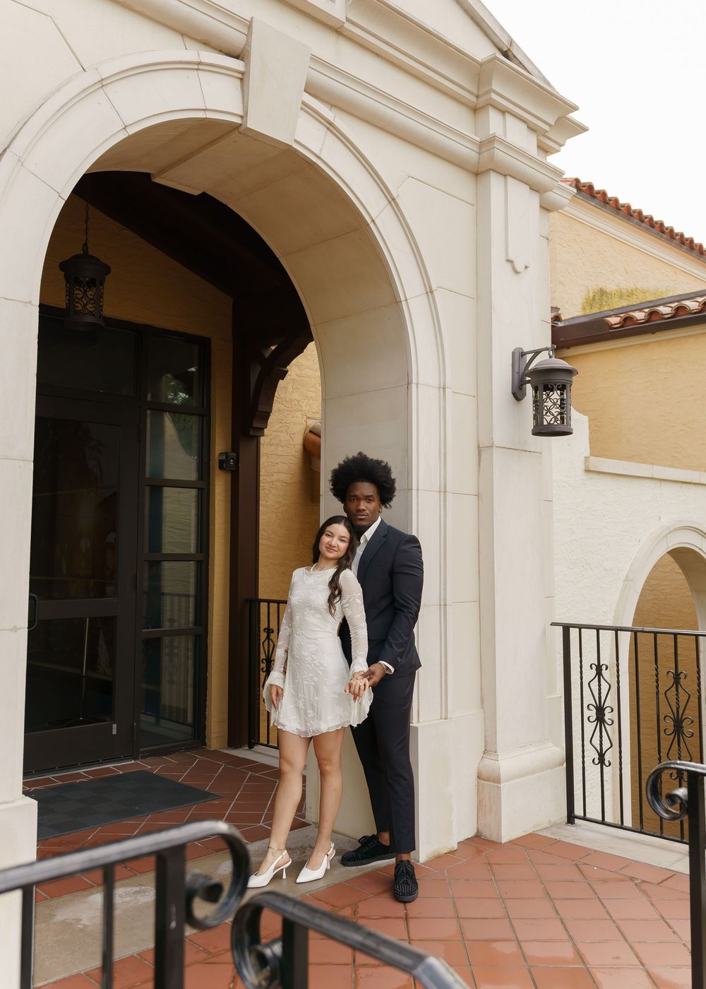 Formal couple stands under elegant white archways on a tiled patio.