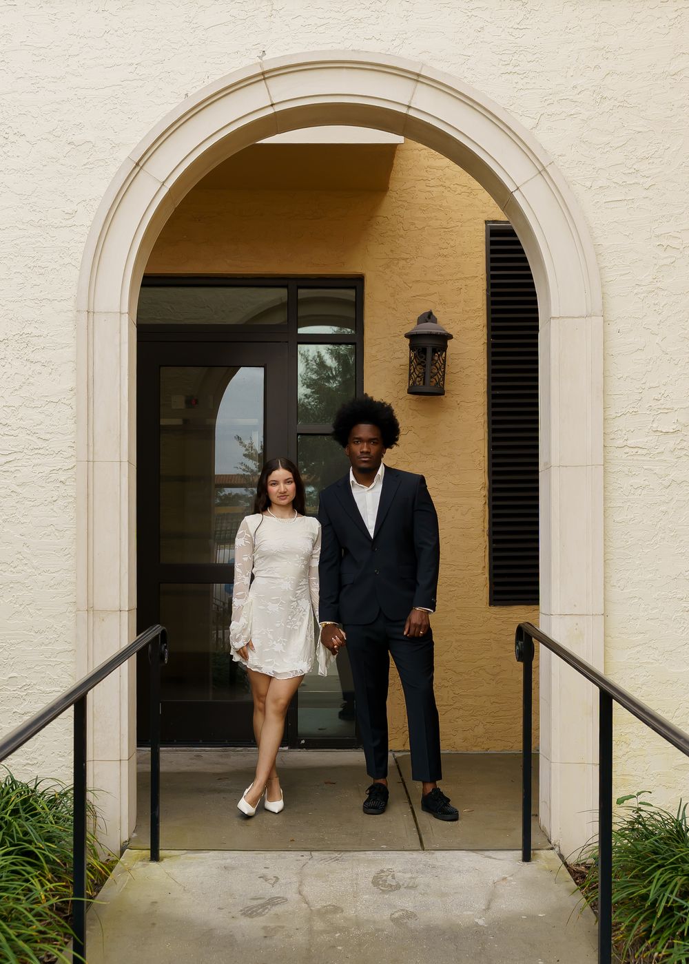 A couple sits together on outdoor concrete steps of an arched entrance to a white building.