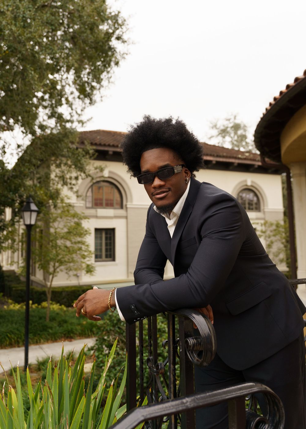 A person in a black suit leans casually against a decorative railing with landscaping and buildings visible.