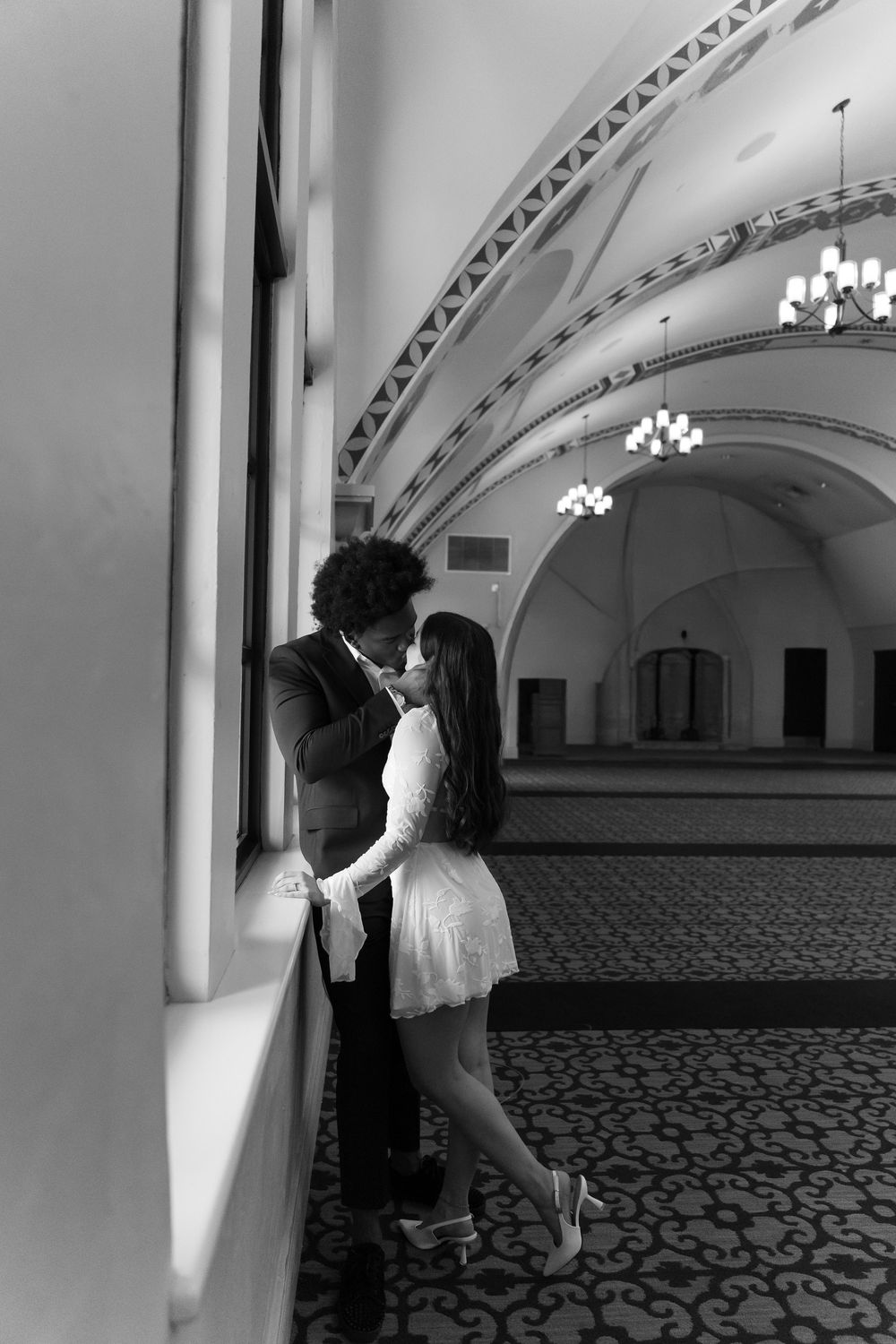 A romantic couple embraces in an elegant hallway with arched ceilings and chandeliers.