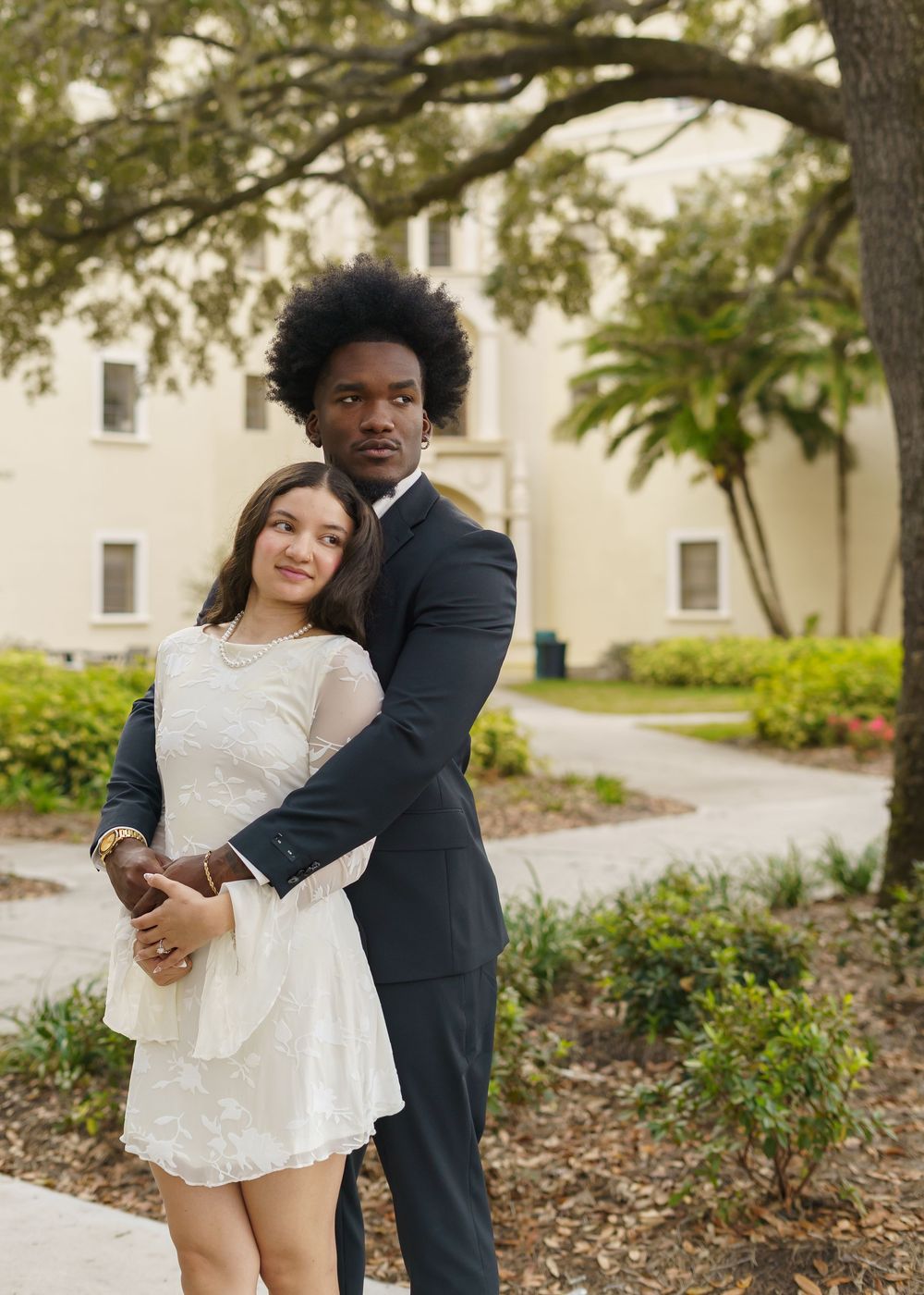 A couple embraces outdoors near historic buildings with lush trees and landscaping in the background.