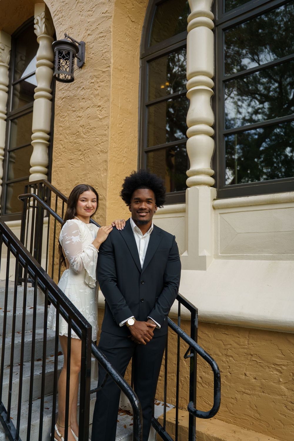 A couple shares playful moments on a porch with white columns and black railings.