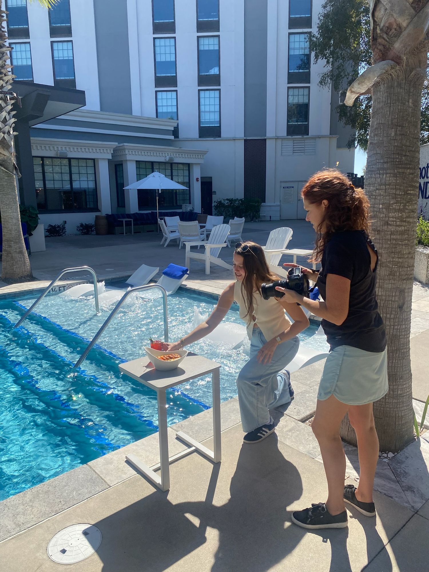 People taking photos of a swimming pool at a modern hotel with palm trees and blue skies in the background.