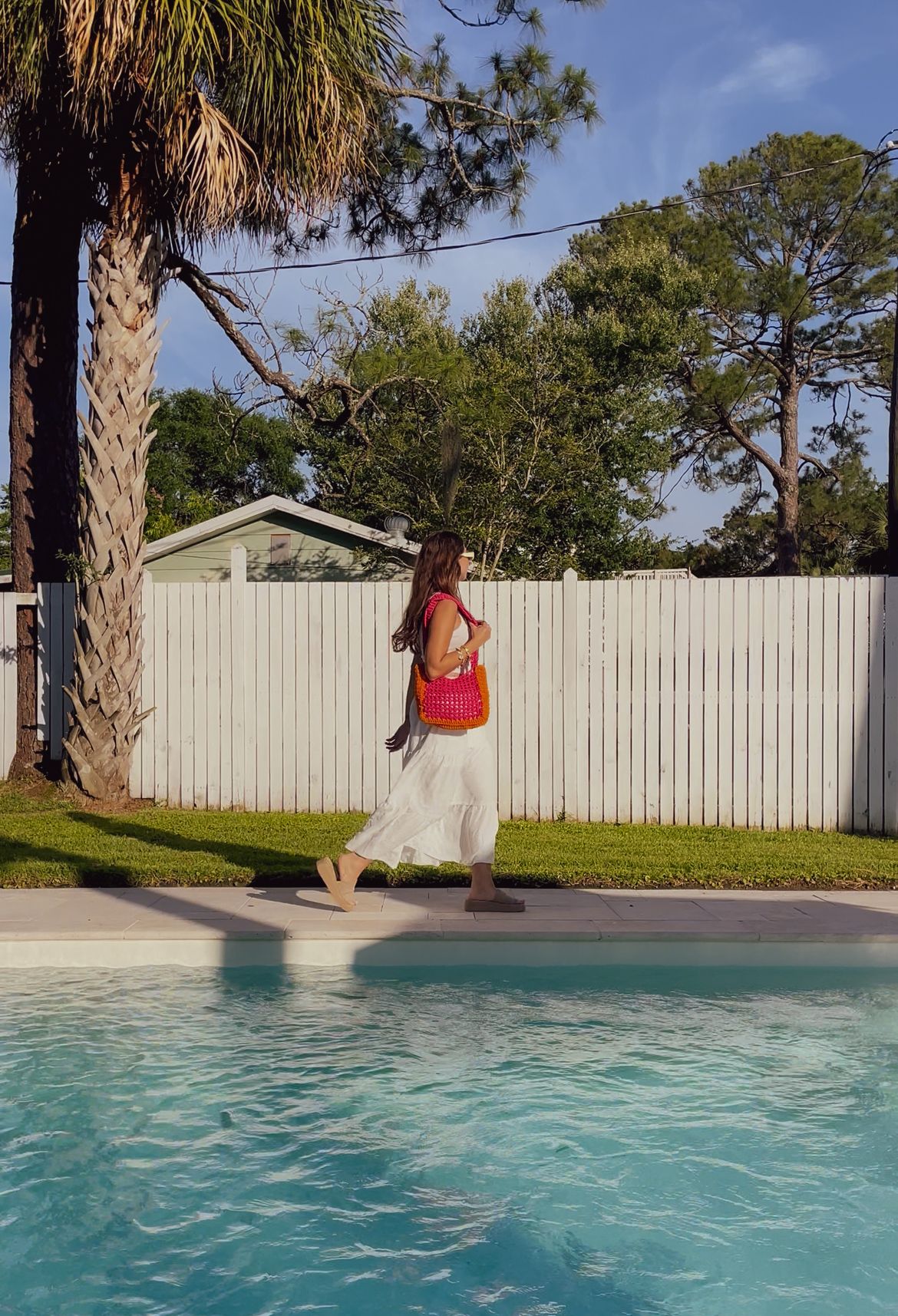 Someone in a pink top and flowing skirt walks along the edge of a swimming pool with palm trees and white fence in background.