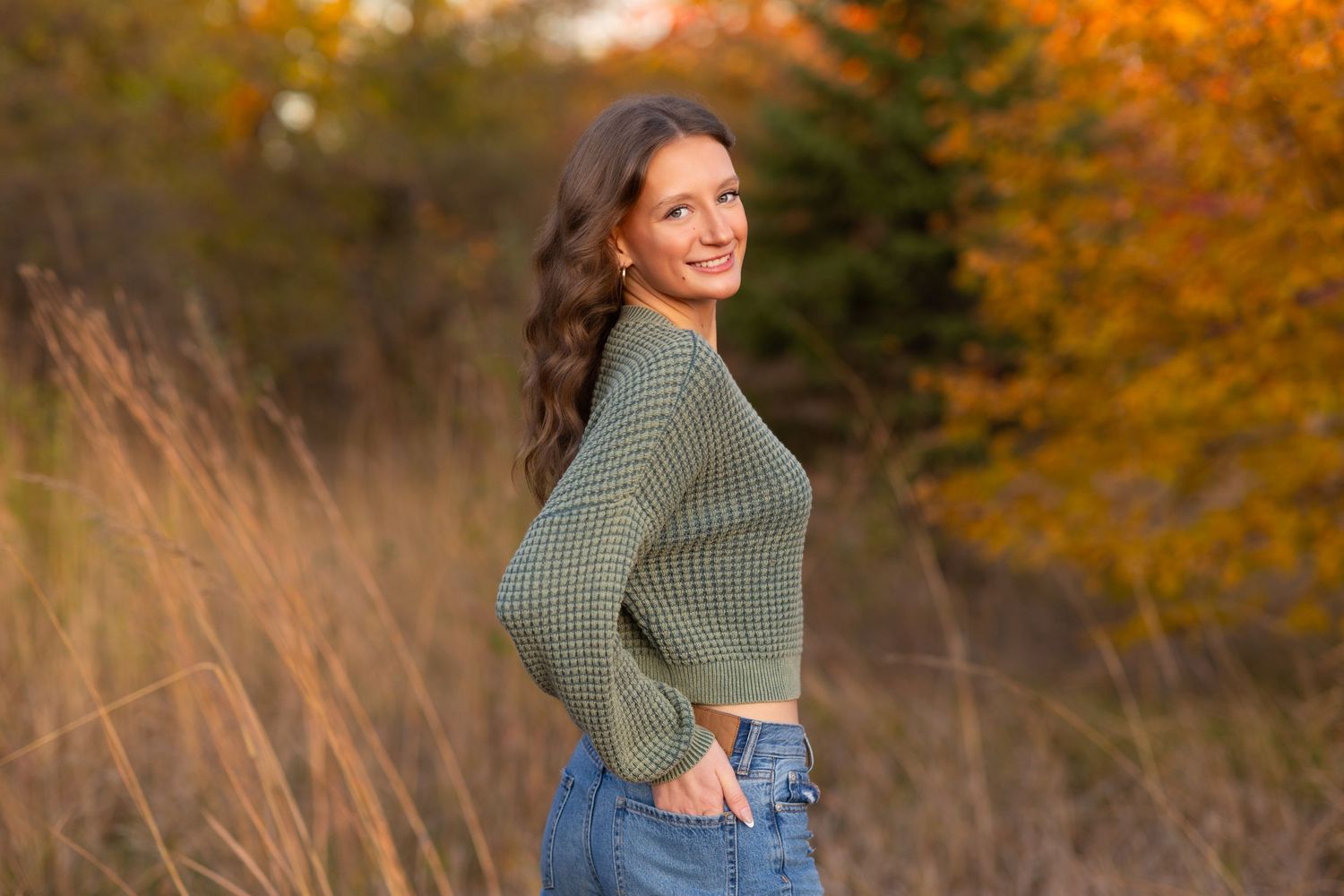 A Senior Session with a girl in a sage green sweater and jeans posing in a field with autumn foliage in the background.