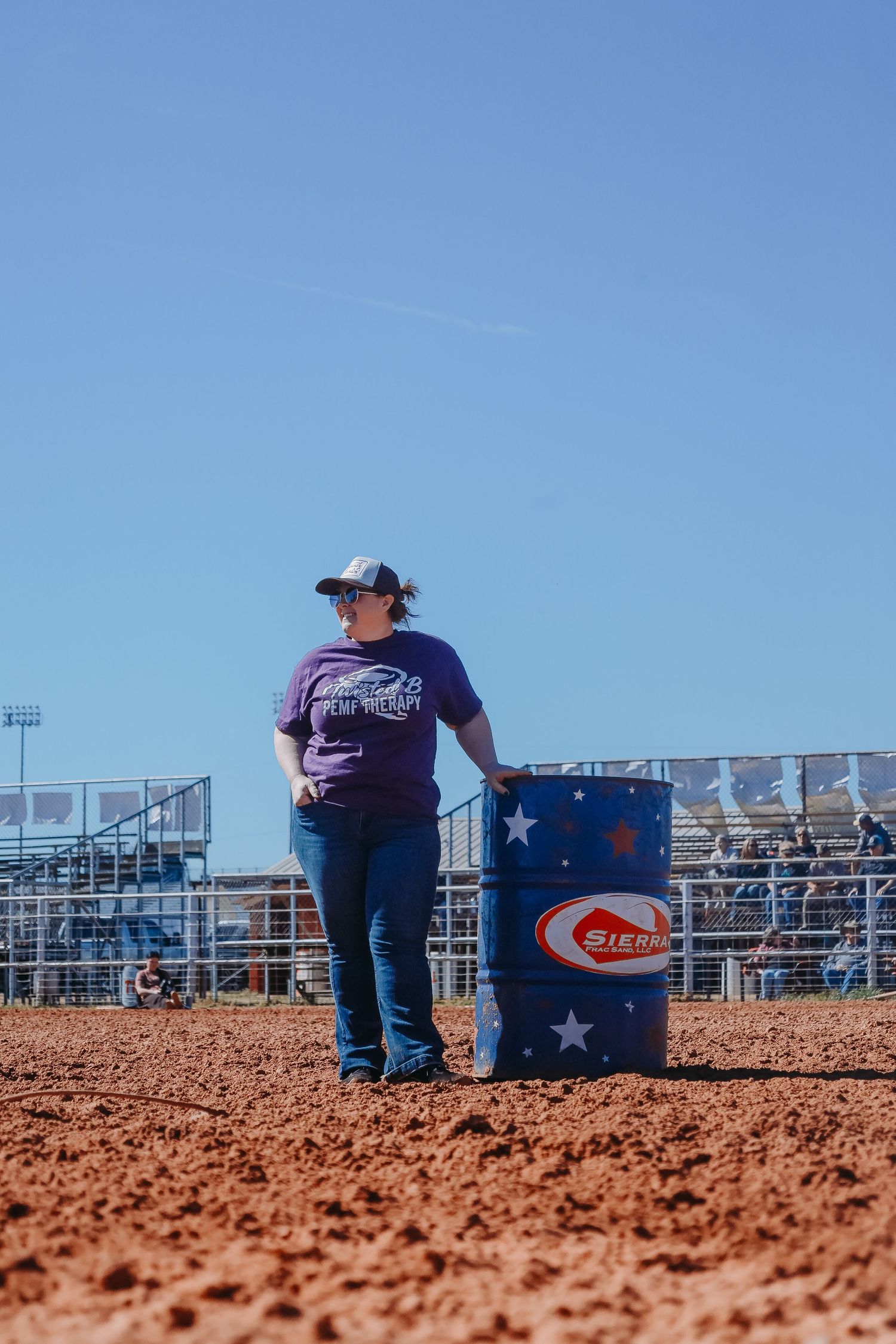 A person stands next to a patriotic blue barrel on red dirt at a rodeo arena under clear blue skies.