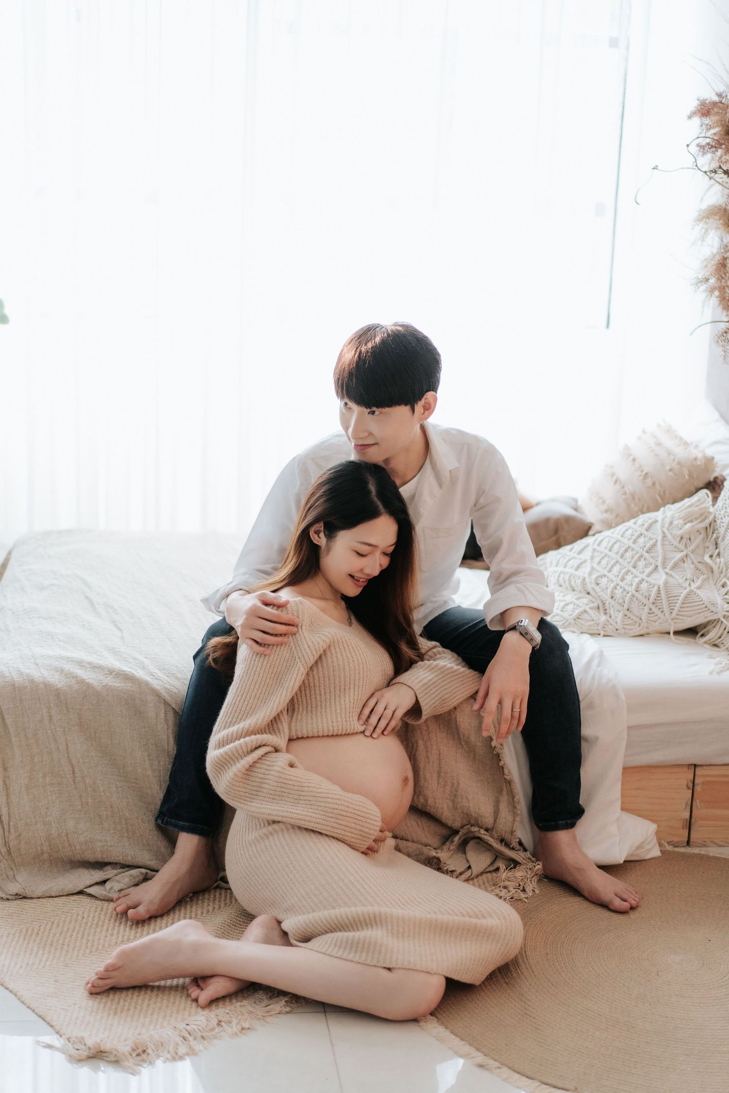 A couple shares an intimate moment on the floor, with the woman wearing a cozy beige sweater and showing her pregnant belly.
