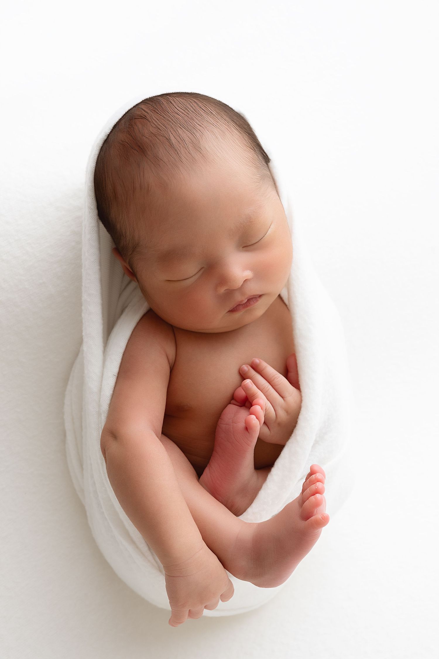 Newborn baby wrapped in white cloth against minimalist white background in artistic portrait style.