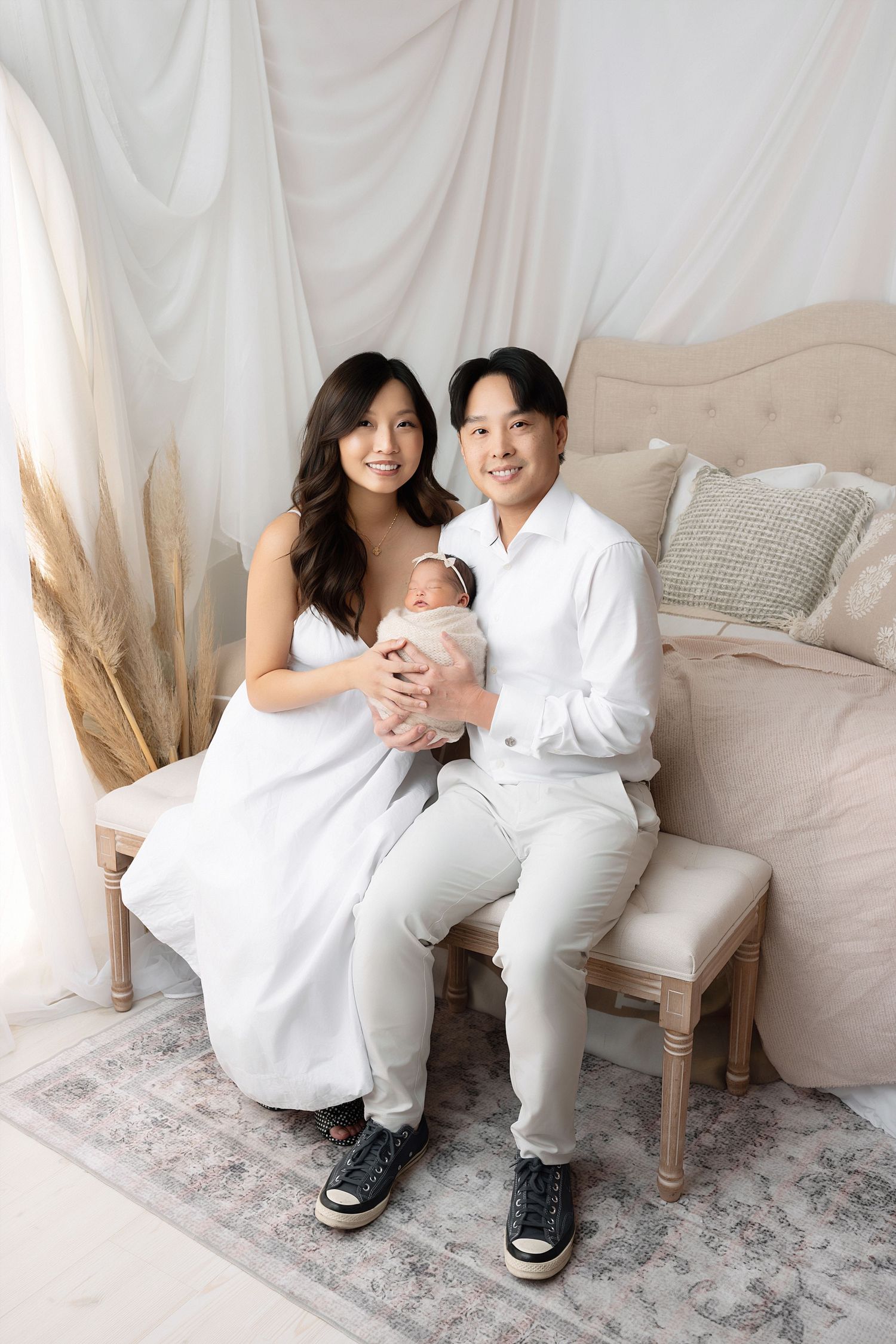 Couple in white outfits sitting together on a beige chair against a light backdrop in a romantic photoshoot setting.