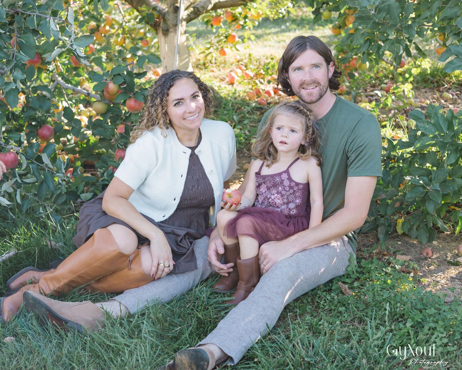 A family sits together on grass beneath an apple tree in an orchard on a sunny day.