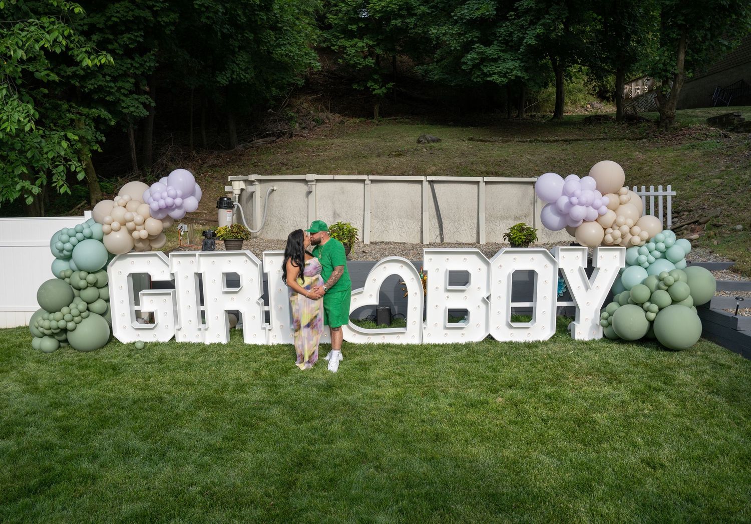 A white balloon arch with green accents decorates an outdoor garden party setup with lettering against a grassy backdrop.