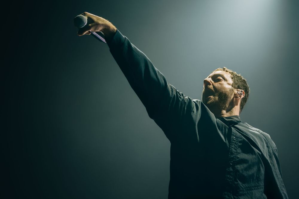 Male singer performing on stage, raising microphone arm under dramatic spotlight in dark concert venue.