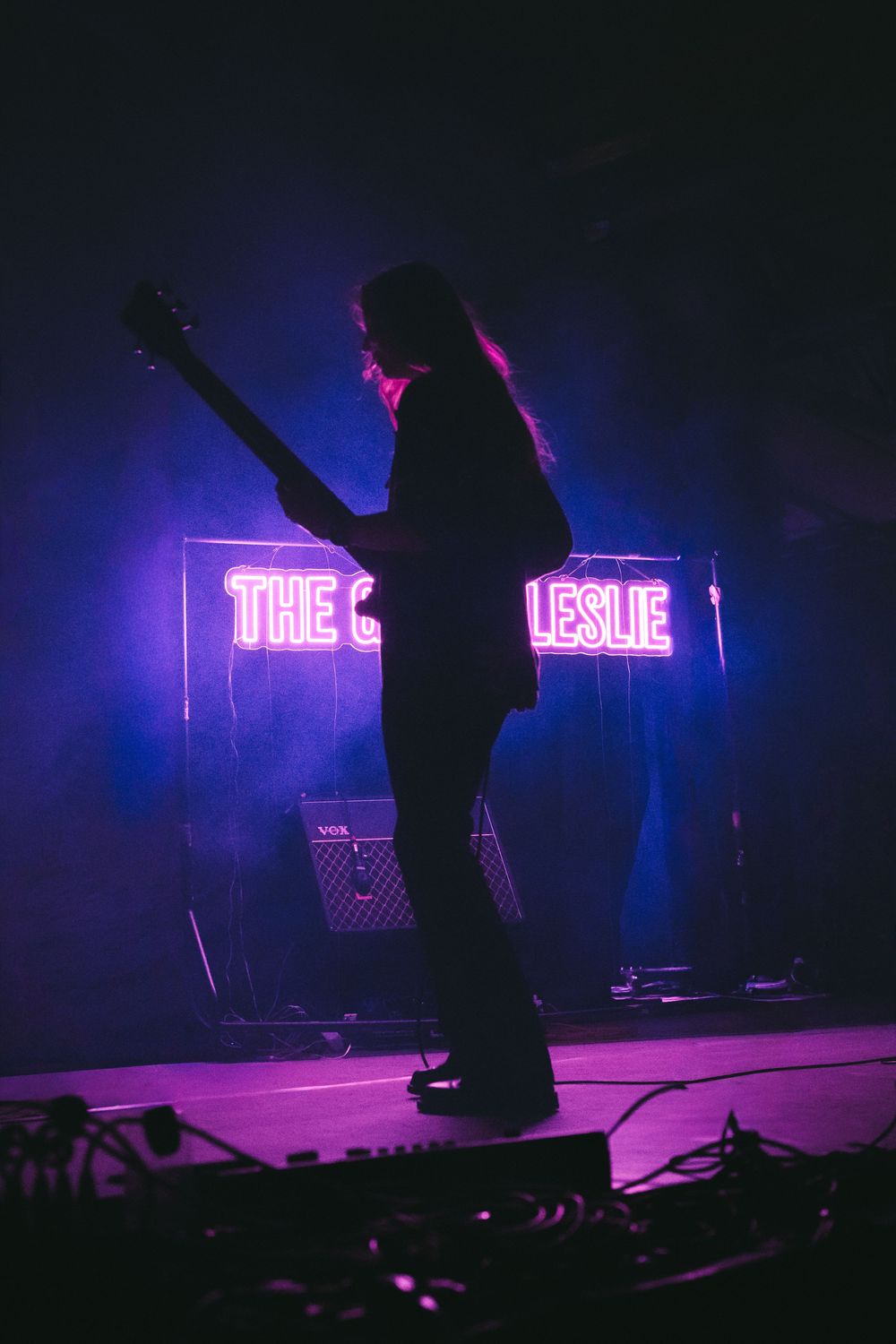 Silhouette of guitarist performing on stage with purple neon sign reading The Leslie glowing in background.