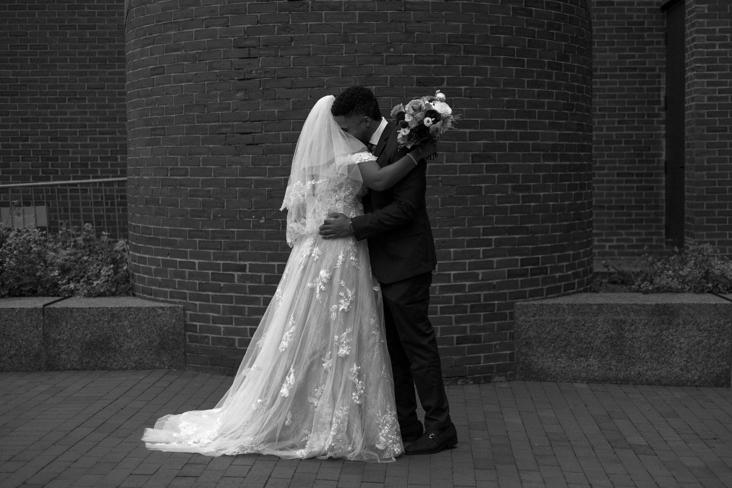 A romantic black and white photo of a couple embracing during their wedding ceremony against a brick wall backdrop.