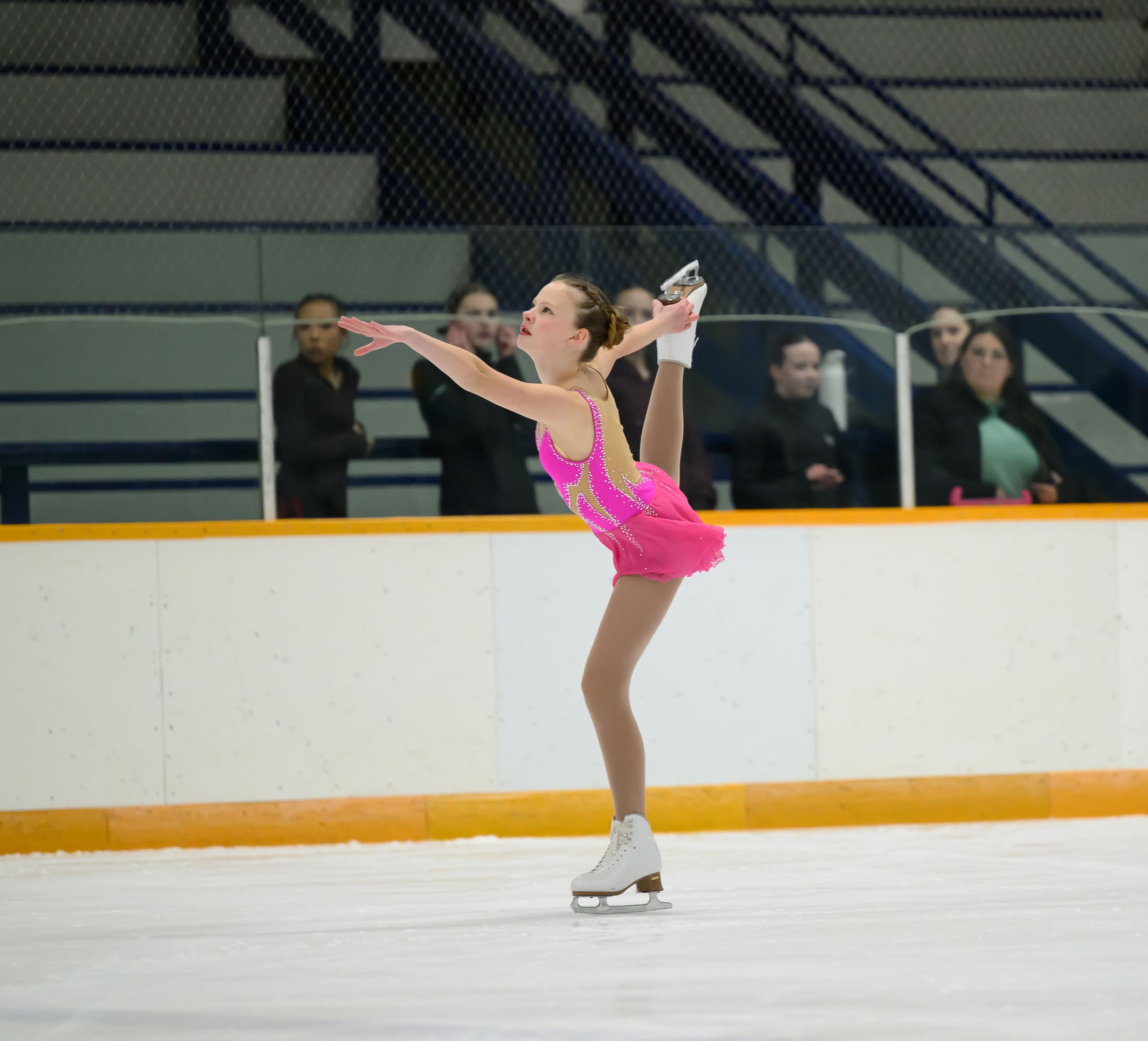 Figure skater in bright pink dress performs graceful pose during competition on ice rink.
