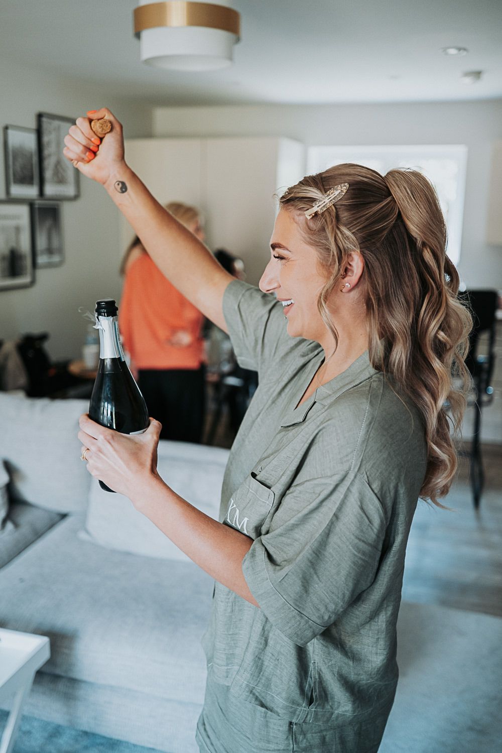 Woman in gray button-up shirt celebrates joyfully with a beverage bottle in hand in a bright modern living room.