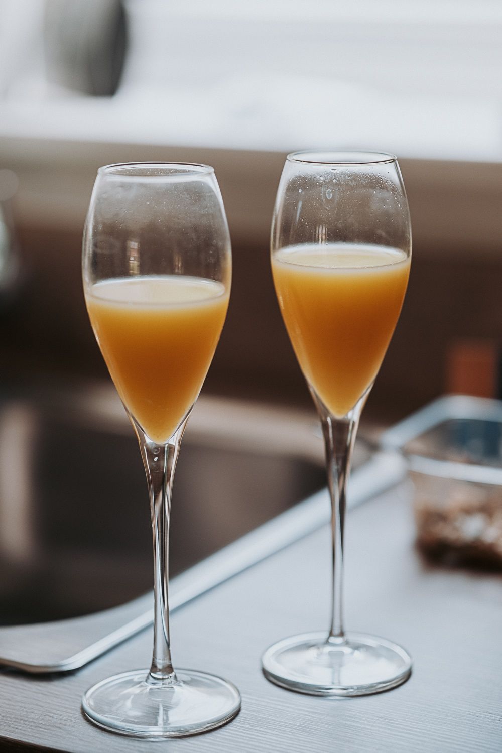 Two elegant champagne flutes filled with orange mimosa cocktails sit on a light colored surface in soft lighting.