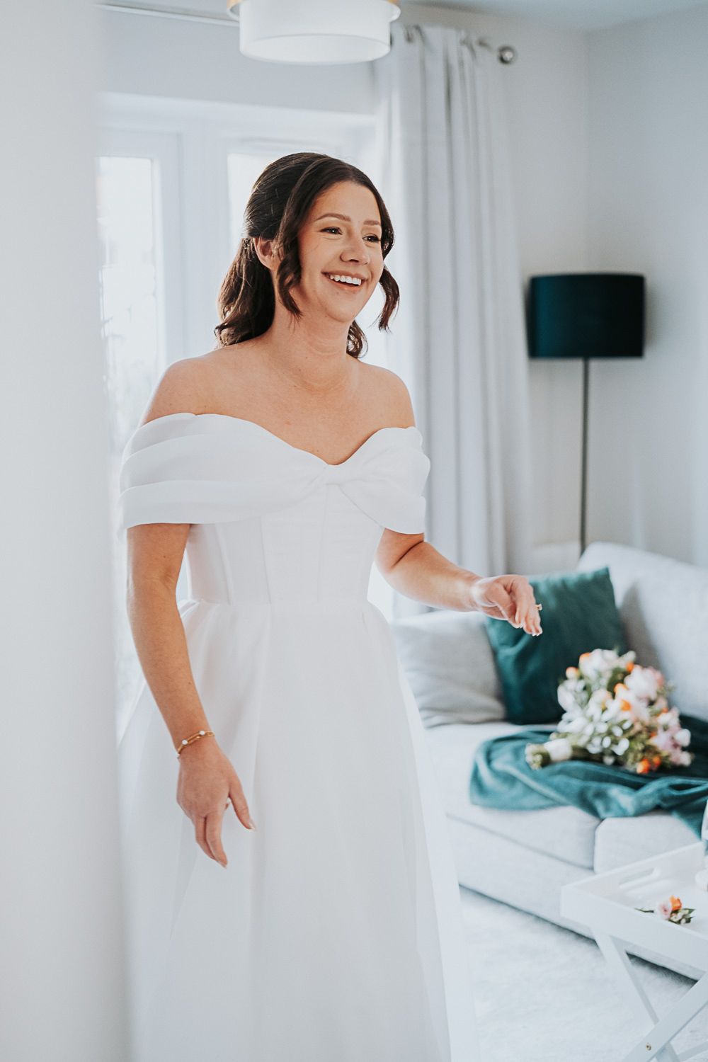 An elegant off-shoulder white wedding dress is worn in a bright modern room with teal accents and floral bouquet.