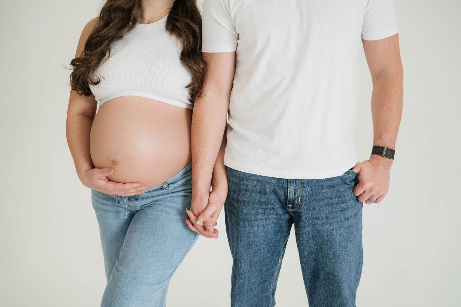Pregnant woman in white shirt holding hands with partner in jeans against white wall.