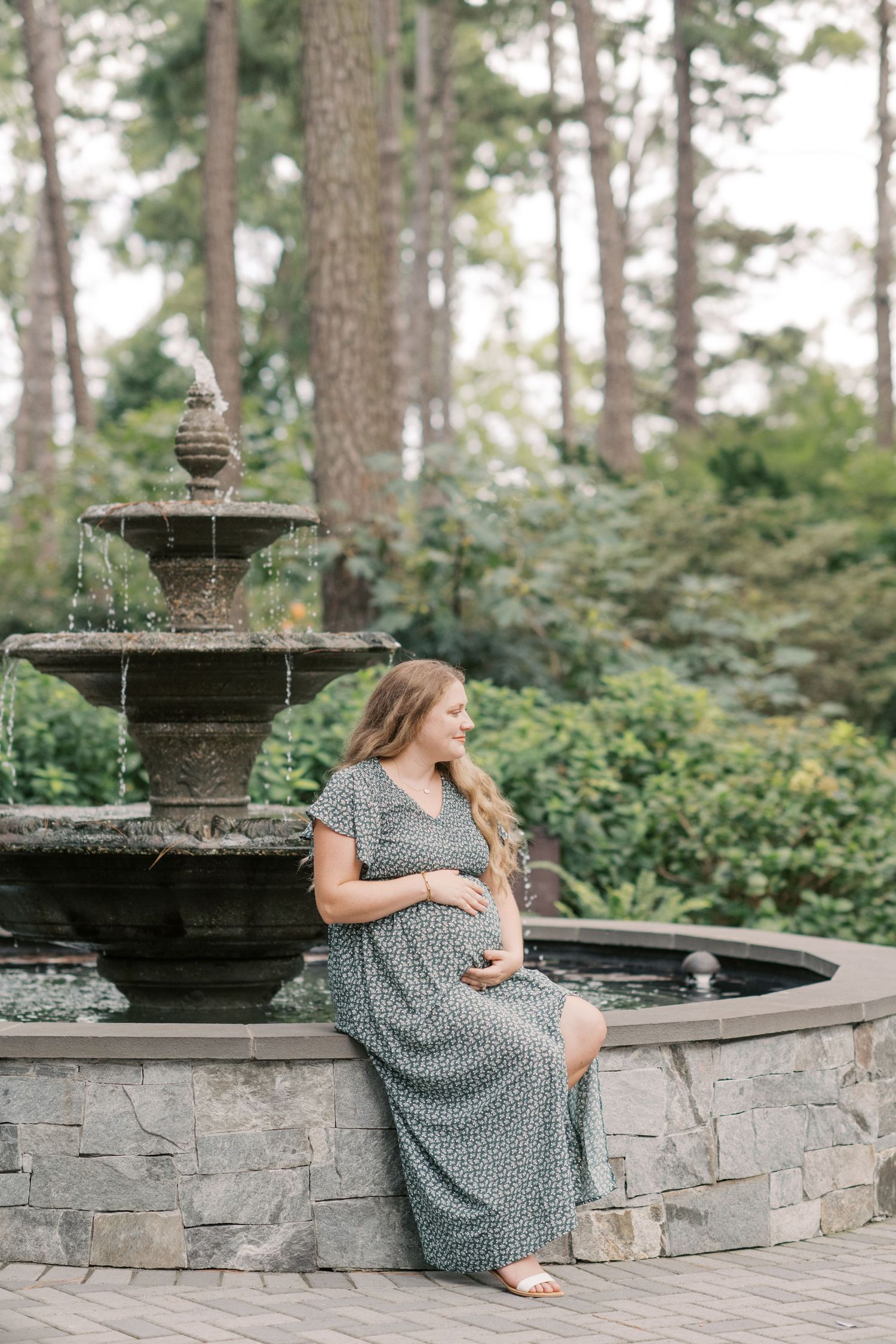 Pregnant woman in floral maxi dress sitting by stone fountain in lush garden, cradling baby bump.