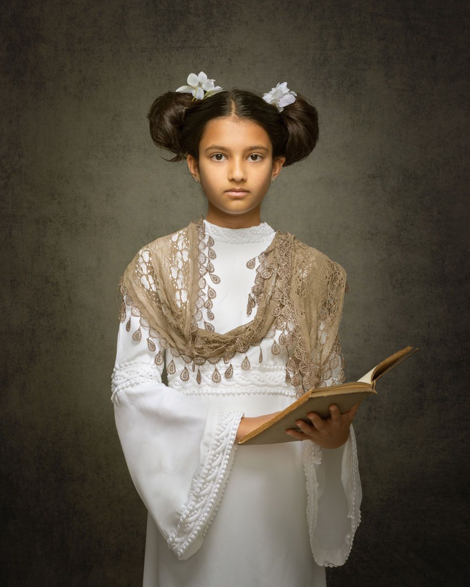Striking fine art portrait of girl, styled in timeless attire and with lavish hairstyle holding an old book