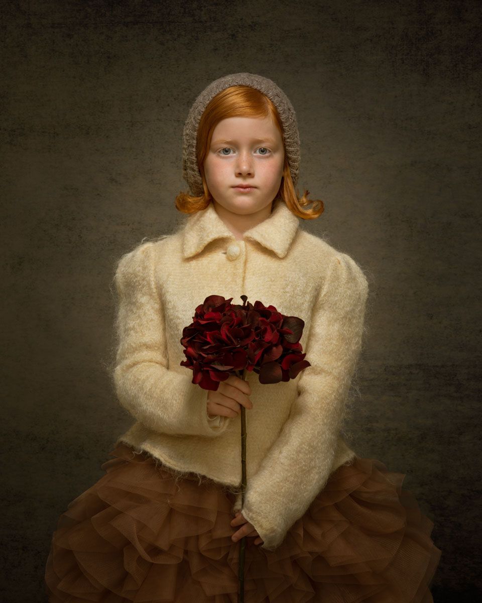 Striking fine art portrait of girl, styled in timeless attire and with lavish hairstyle holding a large rose and stem