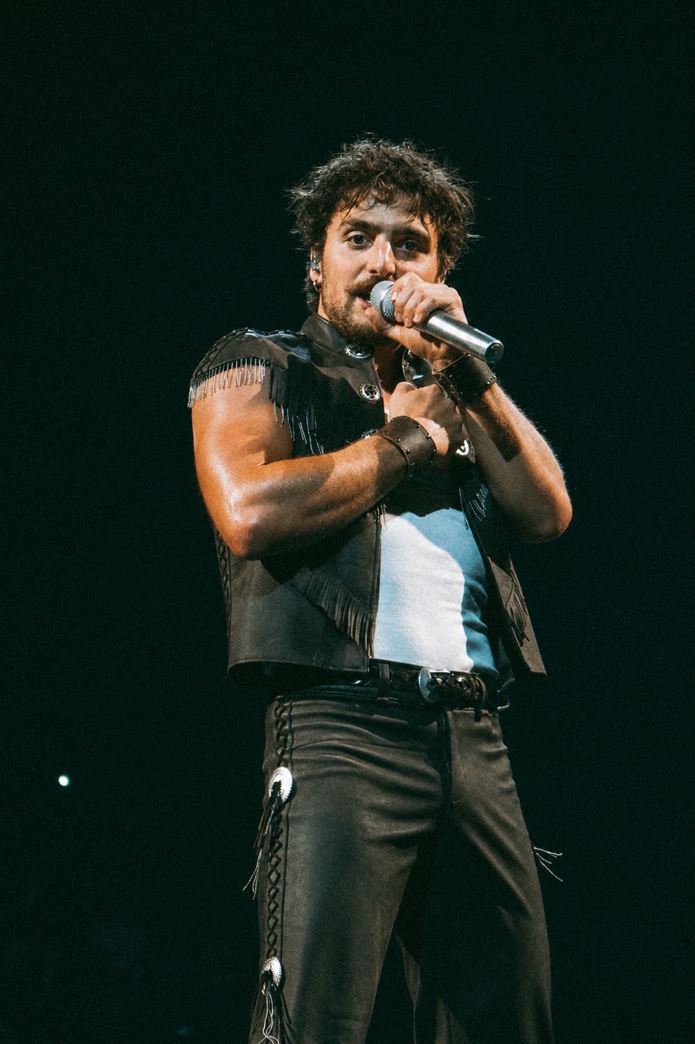 Male rock singer performing on stage, wearing fringe leather vest and black pants, holding microphone in dark concert venue.