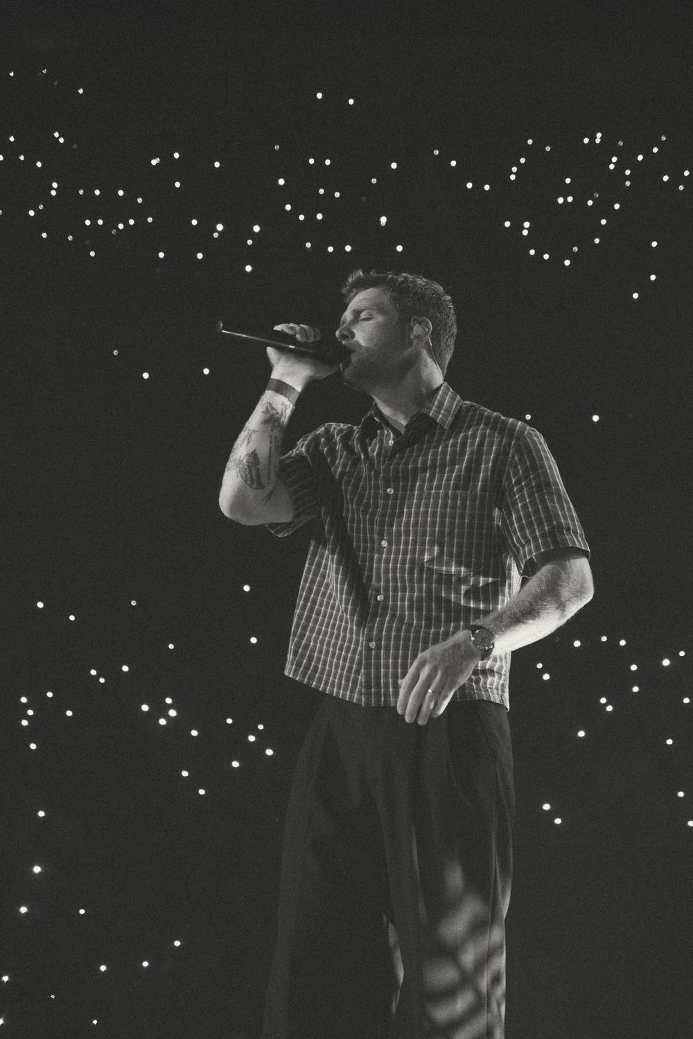 Black and white photo of a male singer performing on stage with microphone, wearing plaid shirt, lights in background.