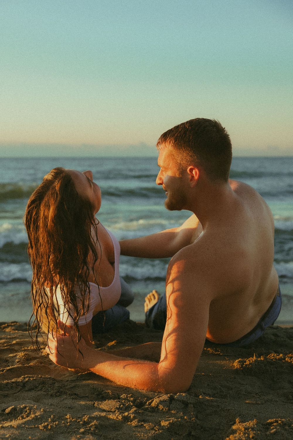 A couple lies together on a sandy beach at sunset as ocean waves roll in along the shoreline.