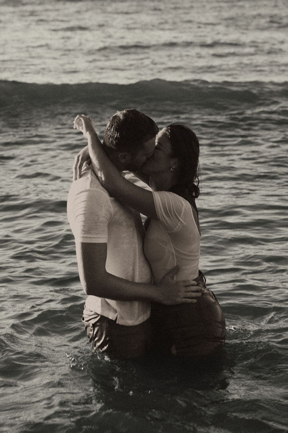 A romantic couple embraces while standing in ocean waves at sunset, creating a moody black and white scene.