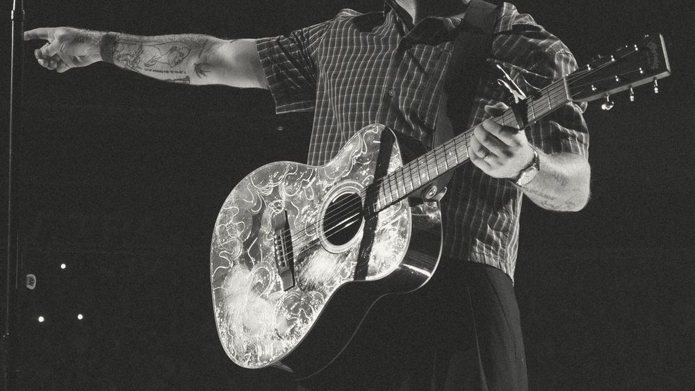 Black and white photo of a male guitarist performing on stage, playing an ornate acoustic guitar while pointing to the crowd.