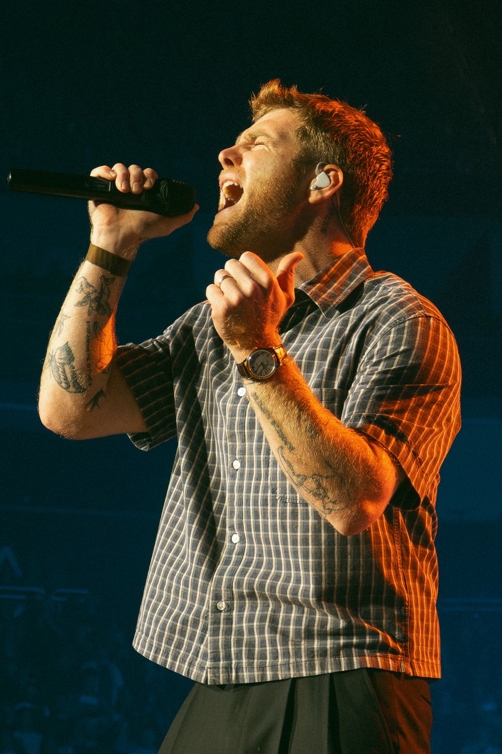 Tattooed male singer performing passionately on stage with microphone, wearing plaid shirt and watch under warm stage lighting.