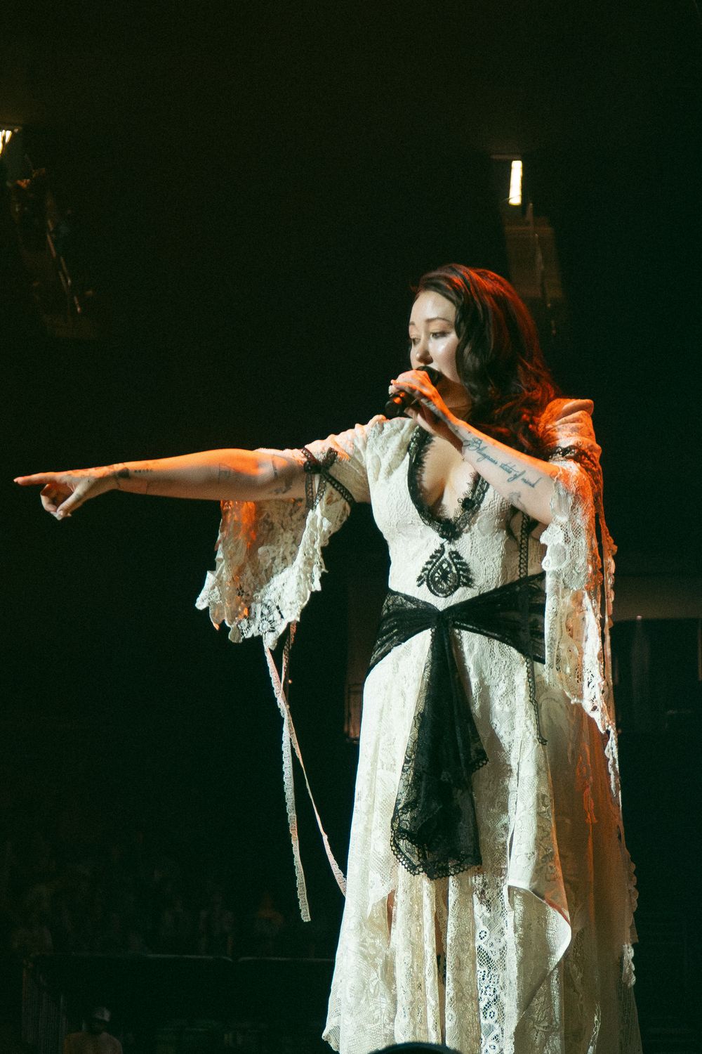 Female singer in white lace gown with black sash performs on dark stage, pointing dramatically while holding microphone.