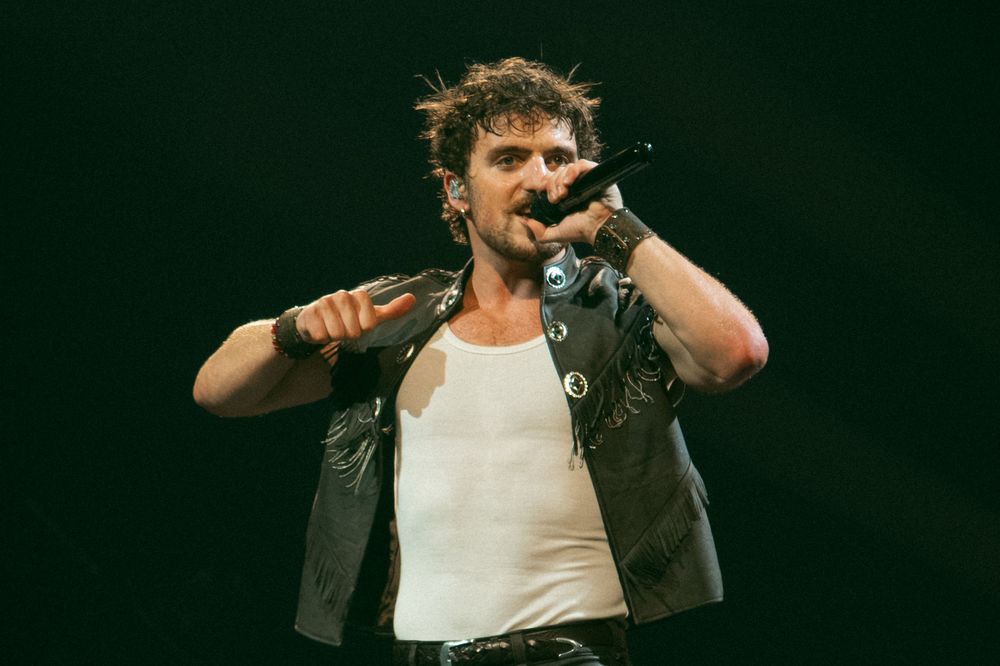 Male rock singer performing on stage, wearing white tank top and leather vest, holding microphone with dramatic expression.