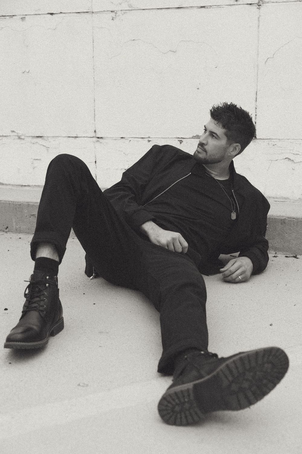 Black and white photo of a man in all-black outfit and boots sitting relaxed on the ground against a white wall.