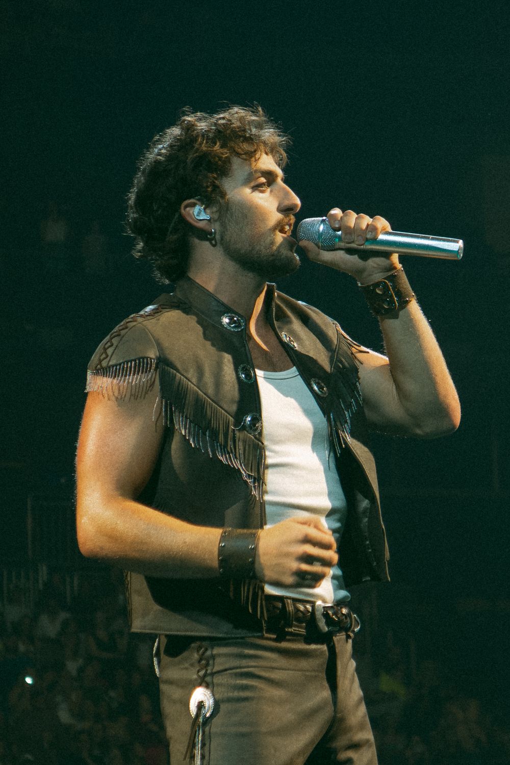 Male singer performing on stage holding microphone, wearing fringed leather vest over white tank top.