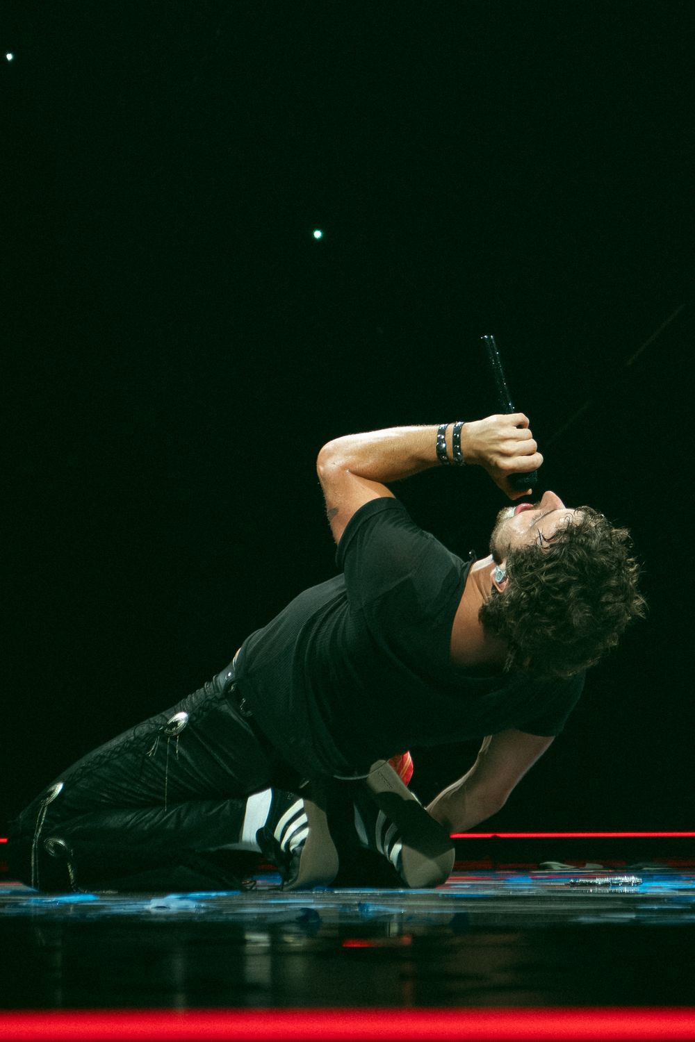 Performer kneeling on stage in black outfit, leaning back dramatically while holding a microphone during a live show.