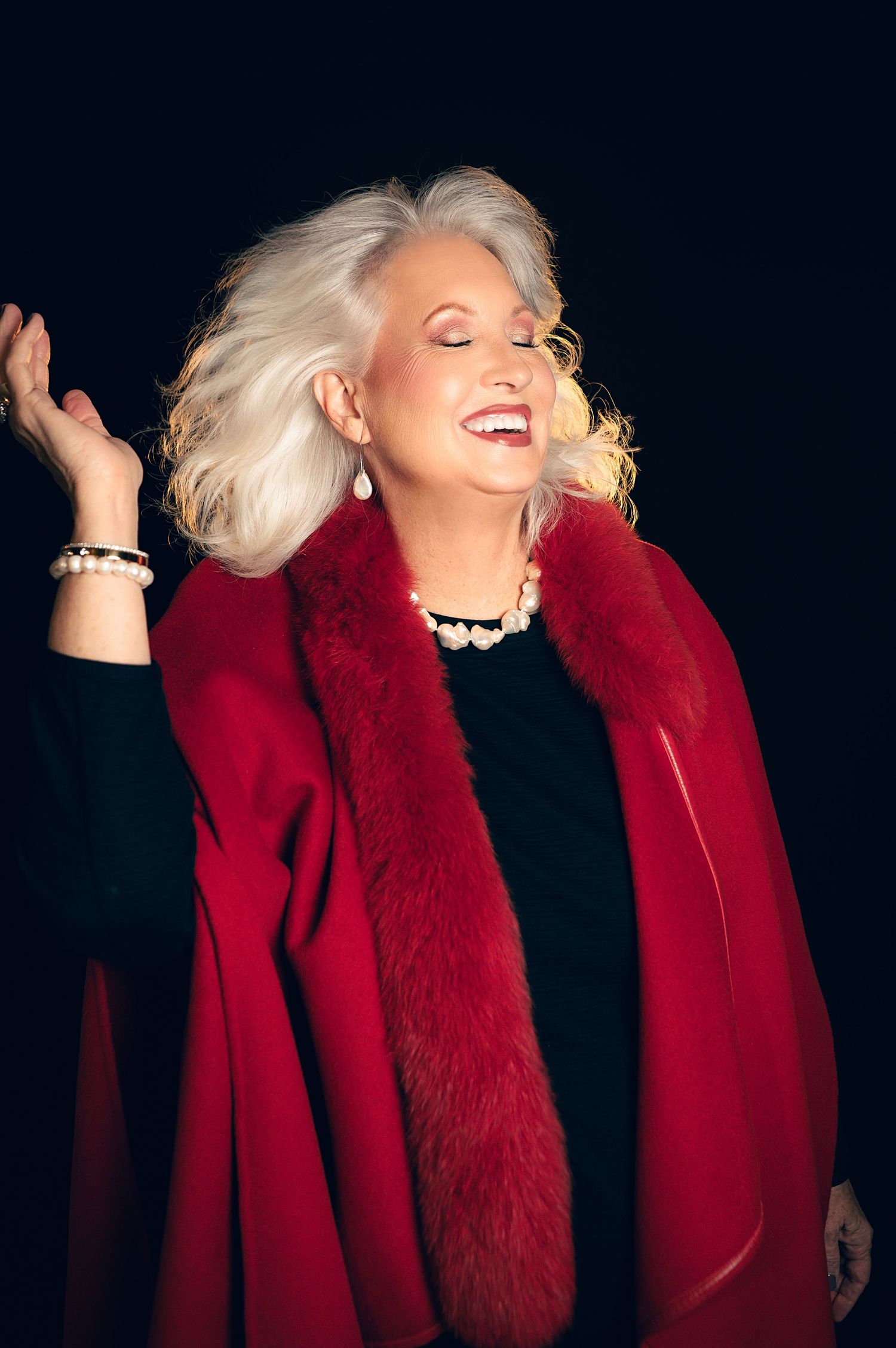 Elegant older woman with white hair laughing joyfully in a red fur-trimmed coat against a black background.