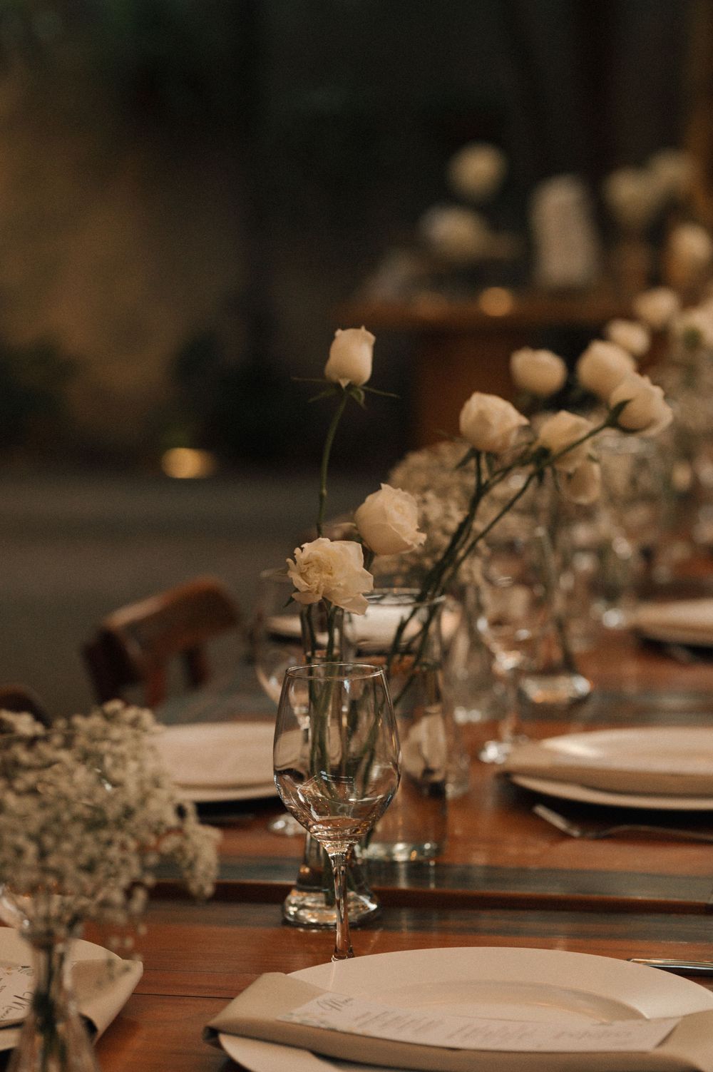 Elegant table setting at a wedding reception with white flowers, wine glasses, and wooden tables in warm lighting.