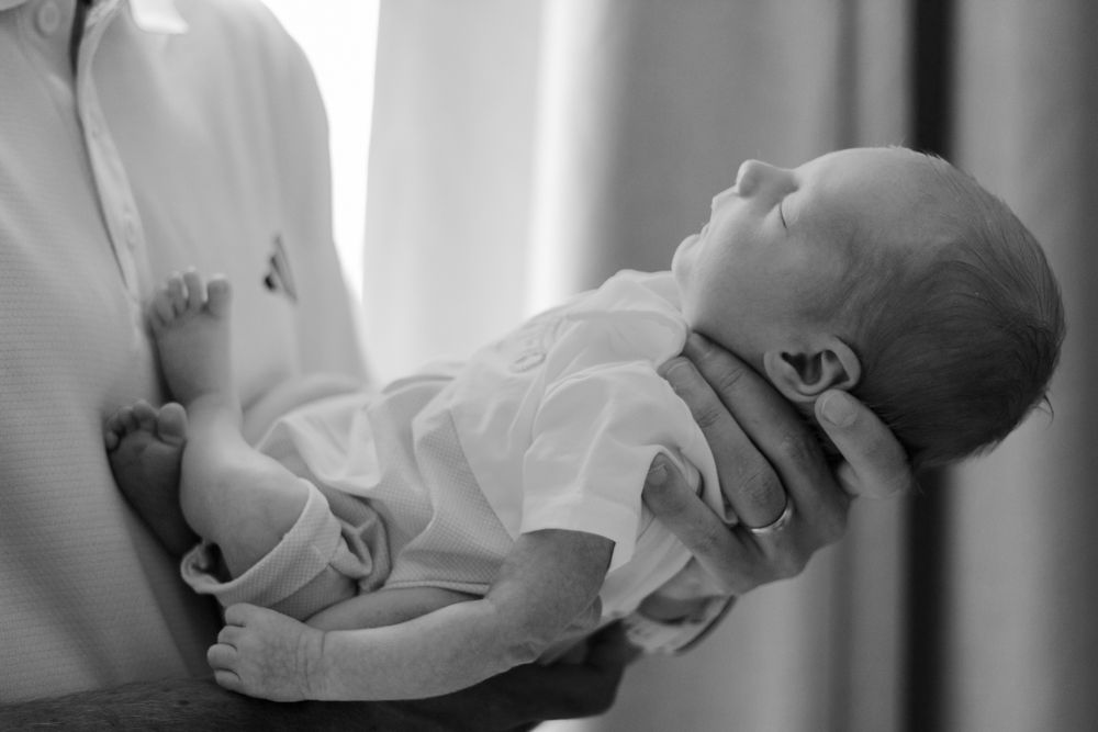 photo of a newborn baby sleeping peacefully in an adult's arms, wearing a white onesie| Westminster, CO motherhood & family photographer