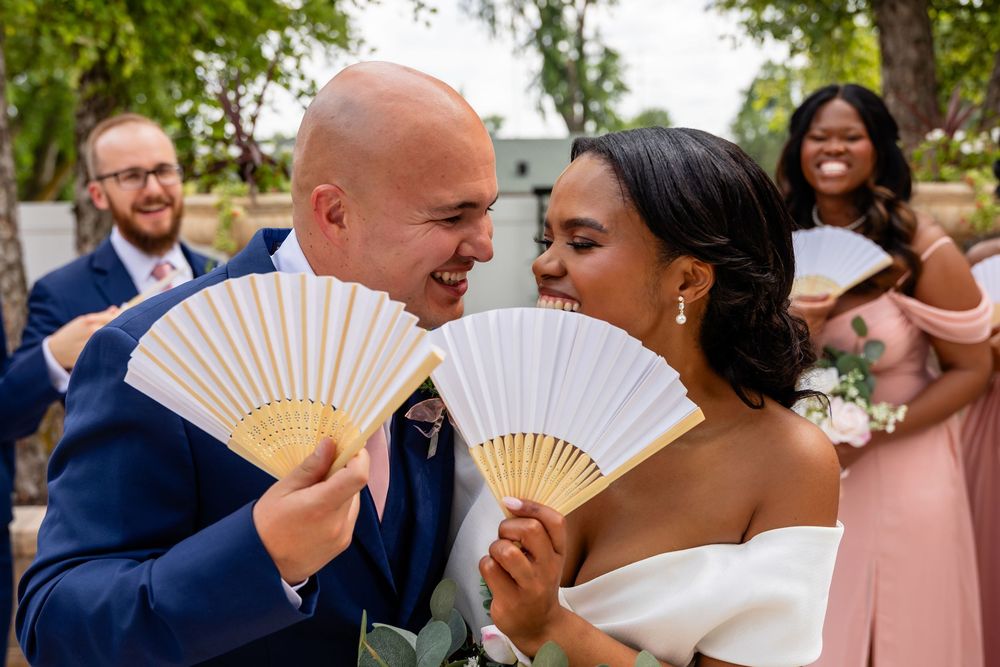 Wedding party shares laughs while holding white fans during outdoor photo session.
