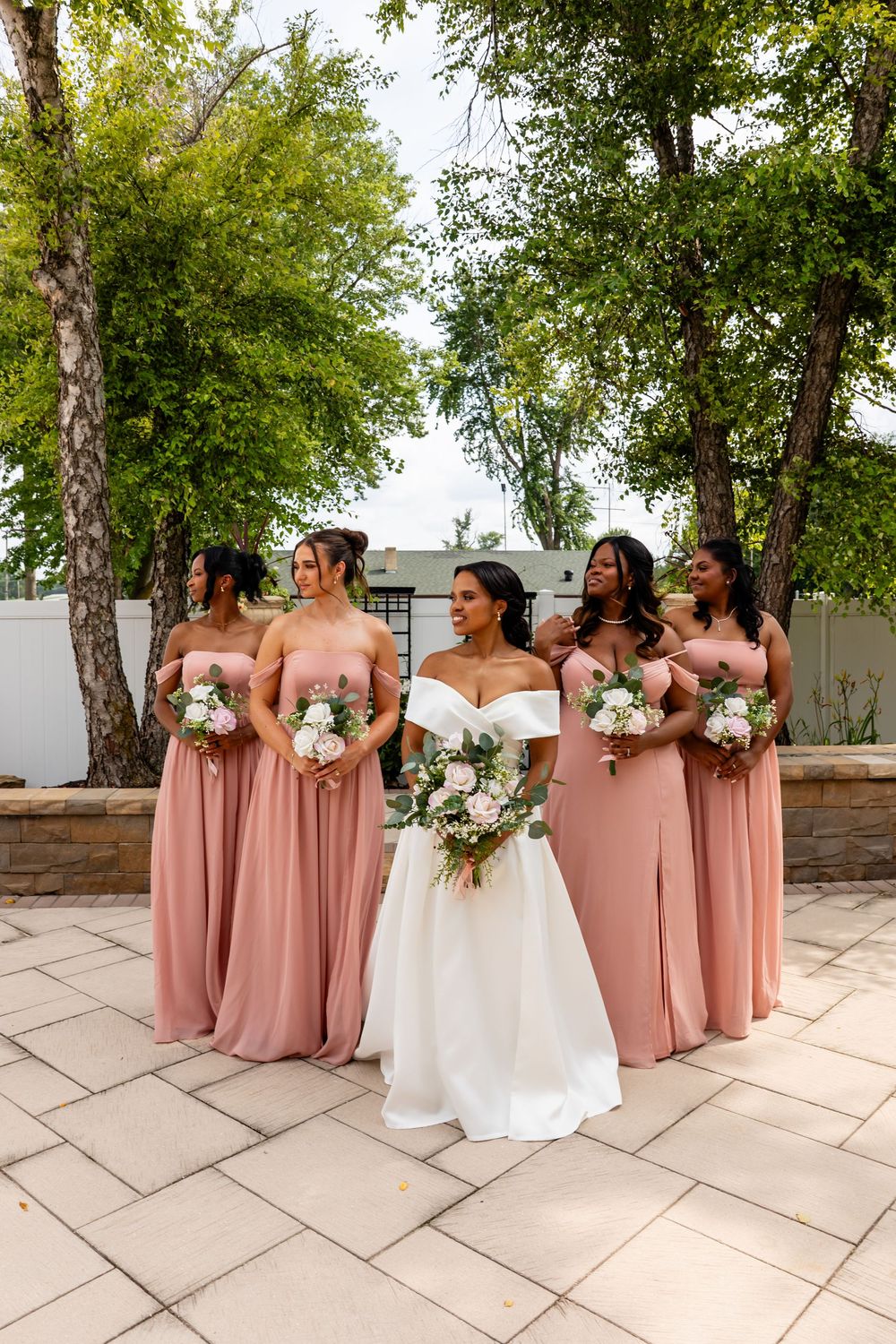 Wedding party poses outdoors with bridesmaids in dusty rose gowns holding white bouquets on stone patio.