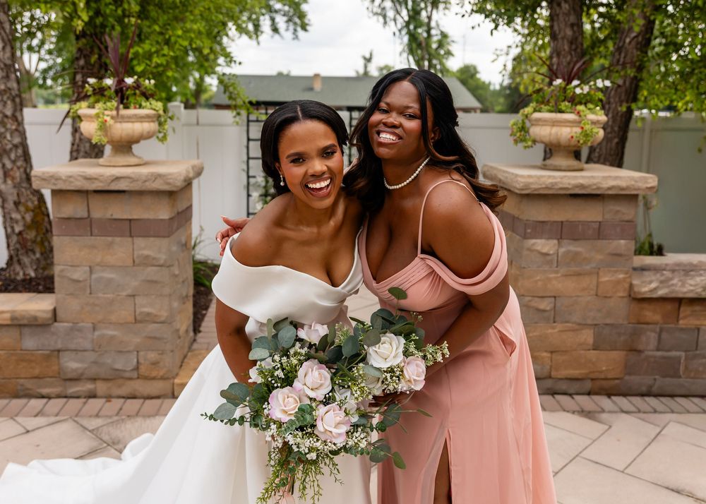 Candid moment between bride in white gown and bridesmaid in pink dress sharing joyful embrace.