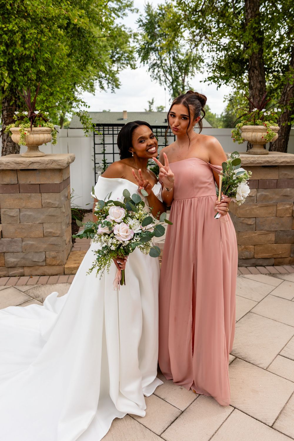 Bride in off-shoulder gown poses with bridesmaid in pink dress against stone wall backdrop.